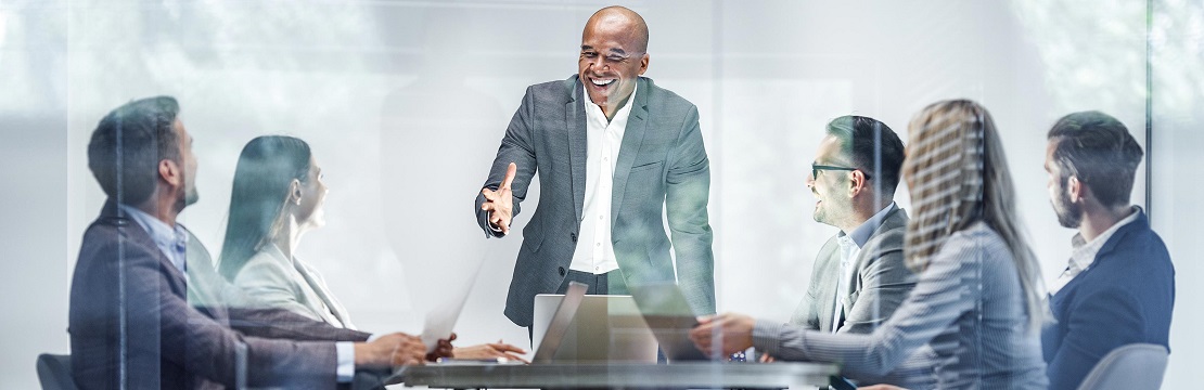 Man stands at the head of a table and is speaking with five individuals seated around the table from him