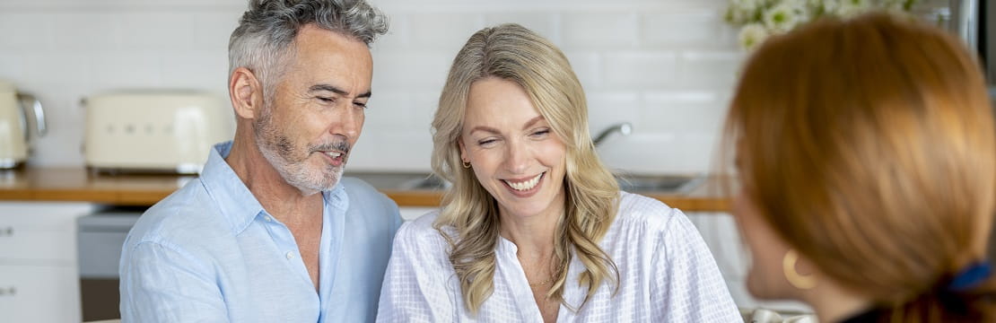 Couple sits at a kitchen table smiling while speaking to an advisor. 